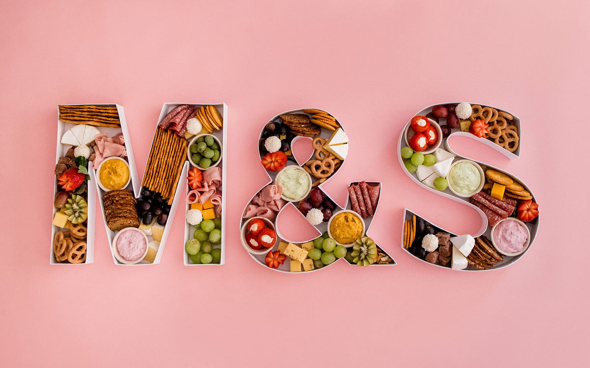 Fresh charcuterie letters filled with cheeses, cured meats, fruit and crackers on a pink background