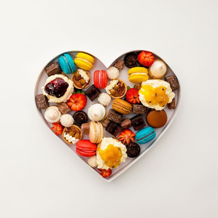 Overhead view of a heart-shaped dessert box with macarons, mini cakes, truffles and strawberries